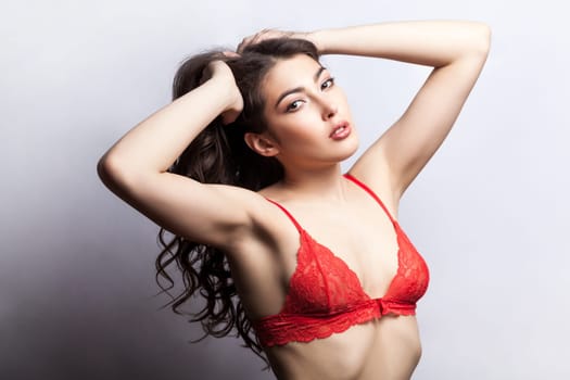 Portrait of attractive brunette woman collecting her wavy hair and looking at camera with passion, raised her arms, wearing red sexy lingerie. Indoor studio shot isolated on gray background.