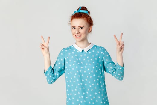 Portrait of positive optimistic red haired woman with hair bun, standing showing v sign, looking at camera with happiness, wearing blue dress. Indoor studio shot isolated on gray background.