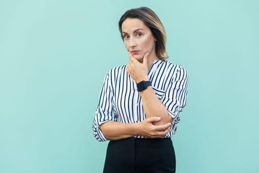 Portrait of thoughtful beautiful middle aged woman wearing striped shirt thinking about solution, looks pensive, looking at camera. Indoor studio shot isolated on light blue background.