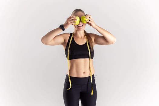 Portrait of funny athletic blonde woman standing with green apples in hands, covering eyes with fresh fruit, wearing black fitness clothing. Indoor studio shot isolated on gray background.