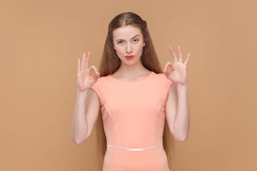 Portrait of adorable positive woman with long hair standing showing okay sign, approval gesture, like service, wearing elegant dress. Indoor studio shot isolated on brown background.