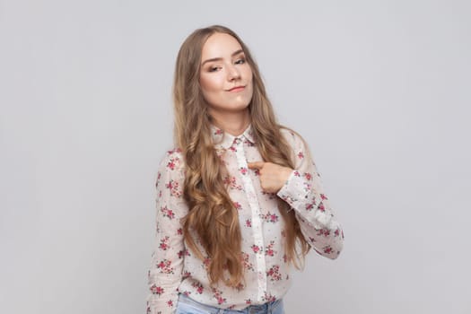 This is me. Portrait of arrogant woman with wavy blond hair pointing at herself, looking egoistic and haughty, proud of achievement. Indoor studio shot isolated on gray background.