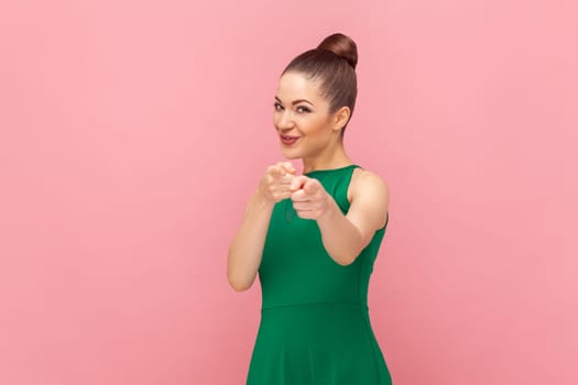 Portrait of smiling satisfied flirting woman standing and pointing to camera with positive expression, need you, wearing green dress. Indoor studio shot isolated on pink background.