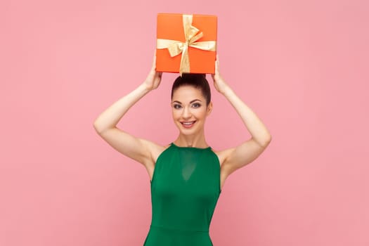 Portrait of satisfied smiling optimistic woman standing holding red present box above her head, being in good festive mood, wearing green dress. Indoor studio shot isolated on pink background.