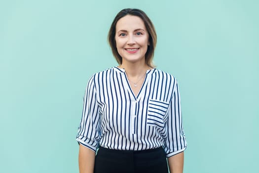 Portrait of smiling positive middle aged woman wearing striped shirt standing and looking at camera, expressing happiness. Indoor studio shot isolated on light blue background.