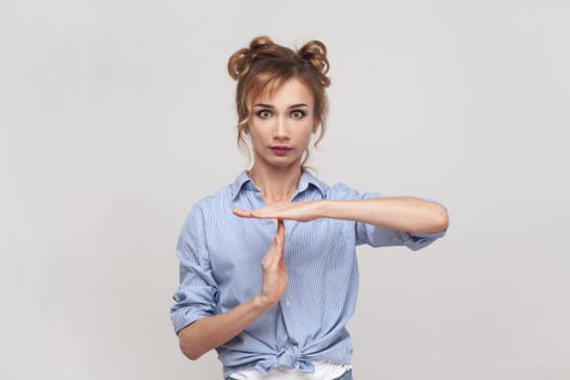 Portrait of blonde woman making pause or break time gesture, timeout sign, asking to wait, upset about deadline, time limit, wearing blue shirt. Indoor studio shot isolated on gray background.