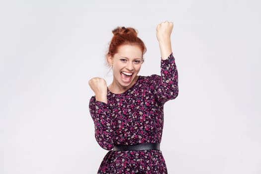 Portrait of ginger woman wearing dress screaming for joy, raising high her fists, celebrating successful winning, incredible victory. Indoor studio shot isolated on gray background.