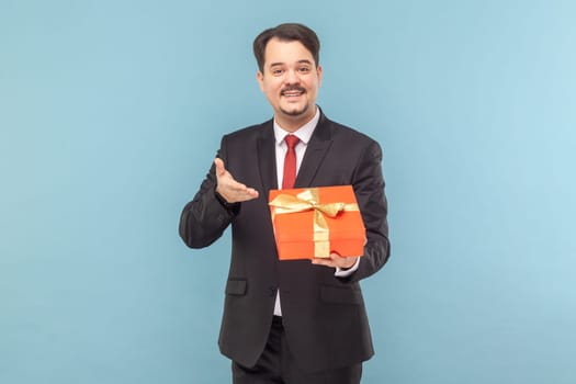 Portrait of handsome smiling happy man with mustache standing showing present box with gift for holiday, wearing black suit with red tie. Indoor studio shot isolated on light blue background.