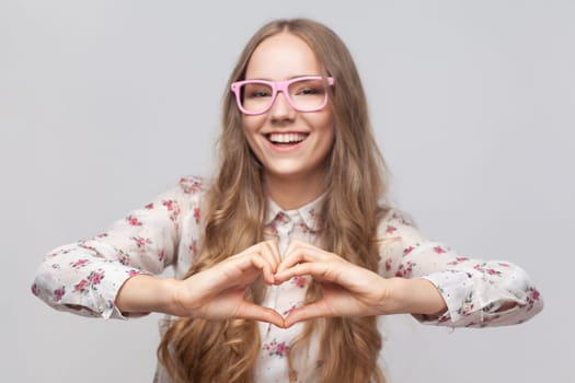 Love you. Portrait of attractive romantic young adult woman in glasses with long blond hair standing making heart with hands, smiling playfully. Indoor studio shot isolated on gray background.