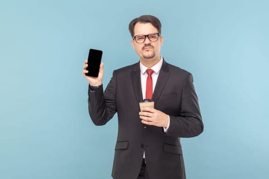 Portrait of sad man with mustache showing smart phone with blank screen, frowning face, drinking coffee to go, wearing black suit with red tie. Indoor studio shot isolated on light blue background.