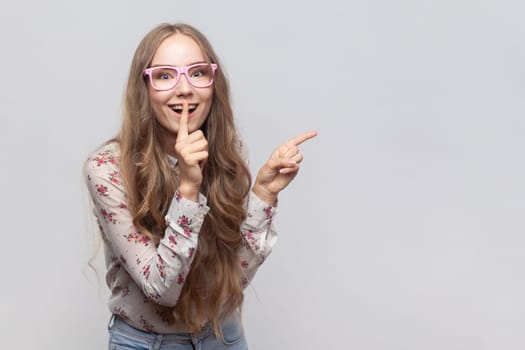 Portrait of positive woman in glasses with long blond hair showing shh gesture holding finger near lips, pointing at advertisement area. Indoor studio shot isolated on gray background.