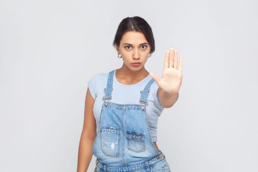 Portrait of serious confident woman wearing denim overalls showing stop gesture with palm of hand, rejecting stress and problems, no discrimination. Indoor studio shot isolated on gray background.