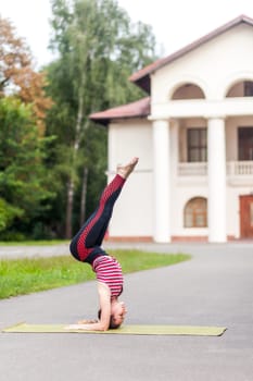 Full length side view of young sporty athletic woman practicing yoga, doing headstand exercise, salamba sirsasana pose, working out outdoor, wearing sportswear, pants and top.