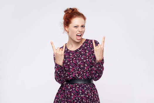 Portrait of crazy ginger woman wearing dress showing rock and roll gesture, heavy metal sign, enjoys favorite music on party, has squints face. Indoor studio shot isolated on gray background.