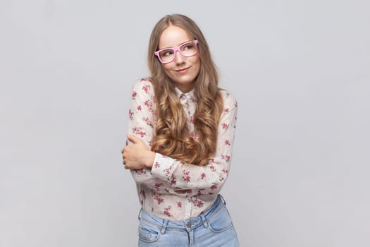 Portrait of woman in glasses with wavy blond hair standing hugging herself and feeling good, appreciating her appearance, self esteem concept. Indoor studio shot isolated on gray background.