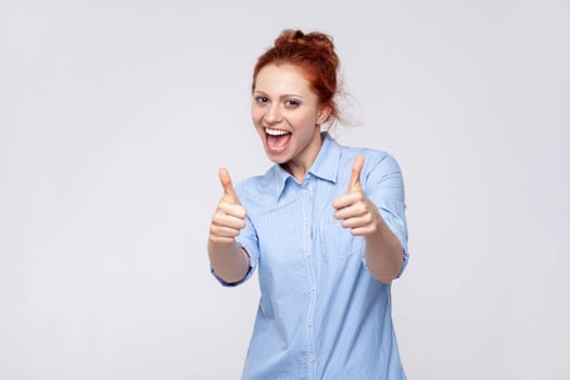 Portrait of joyful cheerful delighted young adult ginger woman wearing blue shirt showing thumbs up, enjoying service, recommend. Indoor studio shot isolated on gray background.