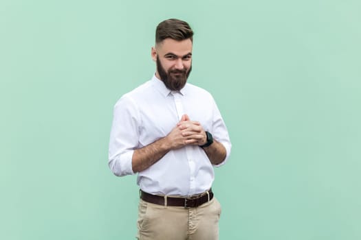 Portrait of young adult bearded businessman wearing white shirt looking at camera with cunning tricky face and smirk, planning evil trick. Indoor studio shot isolated on light green background.