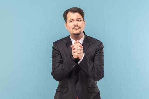 Portrait of sad upset worried man with mustache standing and pleading, looking at camera, asking to forgive him, wearing black suit with red tie. Indoor studio shot isolated on light blue background.