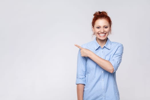 Portrait of ginger woman wearing blue shirt looking at camera and pointing away, presenting copy space for advertisement or promotion. Indoor studio shot isolated on gray background.