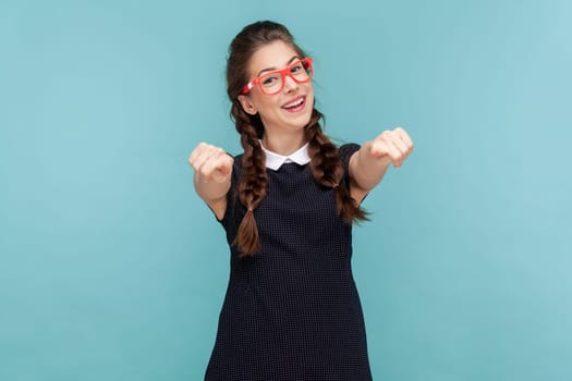 Portrait of smiling cheerful woman with braids pointing at camera indicating at you, making choice, wearing black dress and red glasses. woman Indoor studio shot isolated on blue background