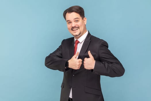 Portrait of happy satisfied man with mustache standing showing thumb up, recommend service, good feedback, wearing black suit with red tie. Indoor studio shot isolated on light blue background.