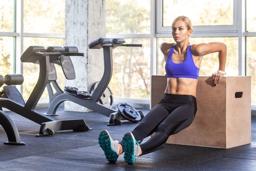 Active blonde sporty woman doing triceps dips using a jump box for her cross training workout routine at the gym, wearing wearing sports top and tights. Indoor shot with window on background.