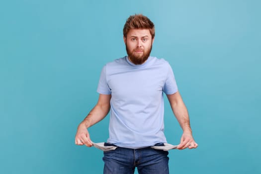 Portrait of sad upset poor bearded man showing empty pockets and looking frustrated about loans and debts, has no money, being jobless. Indoor studio shot isolated on blue background.