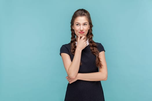 Portrait of cunning woman with braids standing holding chin pondering, thinking about devil plan or prank, wearing black dress. woman Indoor studio shot isolated on blue background.