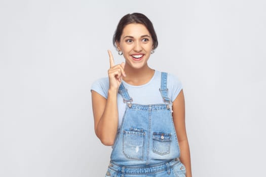 Portrait of inspired excited woman wearing denim overalls pointing finger up and looking amazed about sudden genius idea, got solution. Indoor studio shot isolated on gray background.