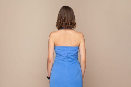 Portrait of unknown woman with wavy hair standing backwards to camera with bare shoulders, wearing elegant blue dress. Indoor studio shot isolated on light brown background.