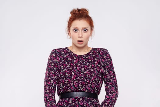 Portrait of young adult ginger woman wearing dress looking at camera with mouth open in amazement, expressing shock, astonishment. Indoor studio shot isolated on gray background.