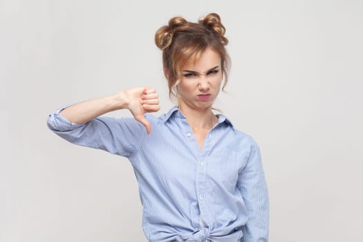 Portrait of disappointed blonde woman shows disapproval sign, keeps thumb down, expresses dislike, frowning face in discontent, wearing blue shirt. Indoor studio shot isolated on gray background.