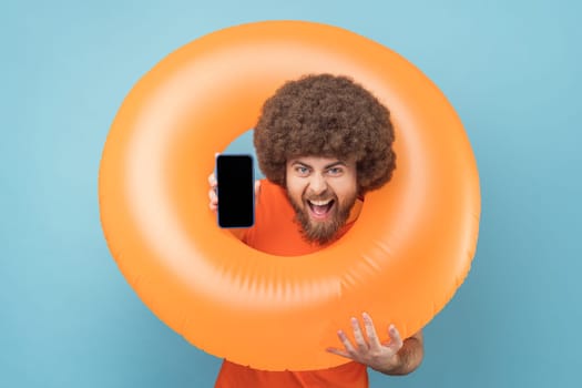 Portrait of man with Afro hairstyle holding orange rubber ring, showing phone with blank screen for advertisement, looking at camera with toothy smile. Indoor studio shot isolated on blue background.