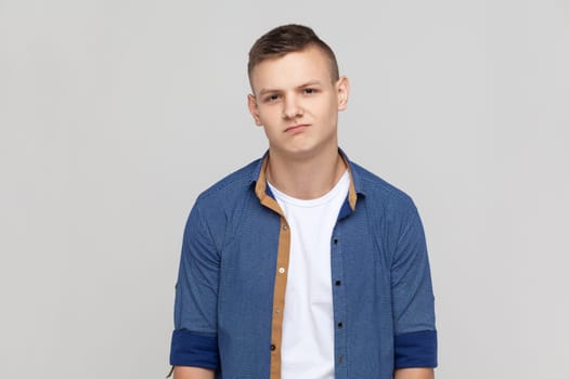 Portrait of depressed sad teenager boy wearing blue shirt being upset of bad news, looking at camera with frowning face, expressing sadness. Indoor studio shot isolated on gray background.