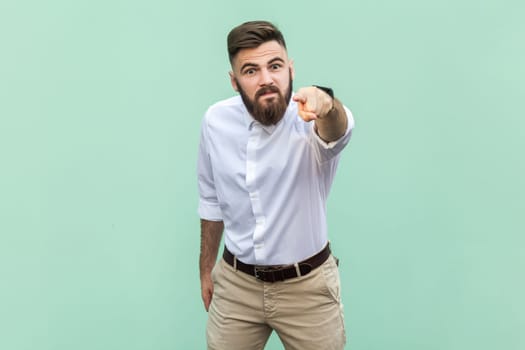 Angry aggressive bearded businessman wearing white shirt looking at camera with serious expression, pointing at you expressing anger and hate. Indoor studio shot isolated on light green background