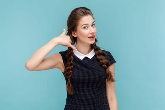 Woman makes phone gesture near, asks boyfriend telephone number, says so call me maybe, has satisfied expression, wearing black dress. woman Indoor studio shot isolated on blue background.