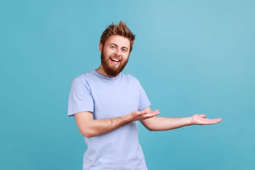 Product advertisement. Portrait of delighted bearded man presenting copy space on palm, showing empty place for commercial text or goods. Indoor studio shot isolated on blue background.