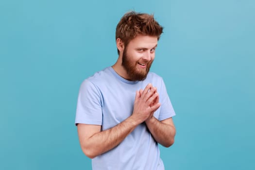 Portrait of cunning bearded man schemes something keeps hands together and makes plans, sly expression looking at camera thoughtfully. Indoor studio shot isolated on blue background.