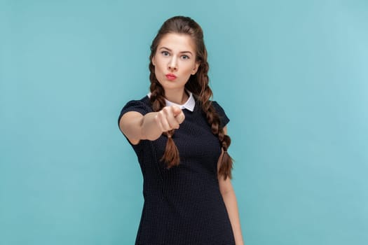 Portrait of bossy strict young woman with braids standing pointing to camera, warning or choosing you, wearing black dress. woman Indoor studio shot isolated on blue background.