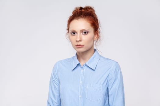 Portrait of sad unhappy upset redhead woman wearing blue shirt standing and looking at camera with dissatisfied sadness face. Indoor studio shot isolated on gray background.