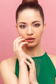 Closeup beauty portrait of beautiful attractive confident woman with bun hairstyle, looking at camera, keeps hand near her lips, wearing green dress. Indoor studio shot isolated on pink background.