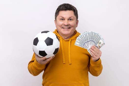 Smiling middle aged man showing soccer ball and fun of hundred dollar bills, winning lot of money betting for sport, wearing urban style hoodie. Indoor studio shot isolated on white background.