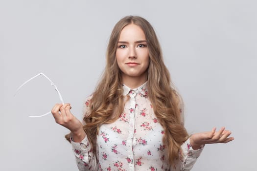 Young adult attractive surprised woman with wavy blond hair standing with raised arms, holding glasses, looking at camera with confused expression. Indoor studio shot isolated on gray background.