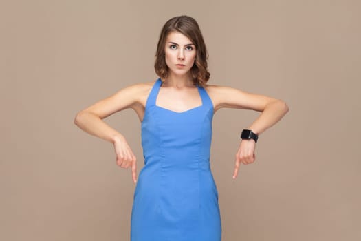Here and right now. Unhappy gloomy woman points down and looks at camera sadly, demonstrates something unpleasant, wearing blue dress. Indoor studio shot isolated on light brown background.