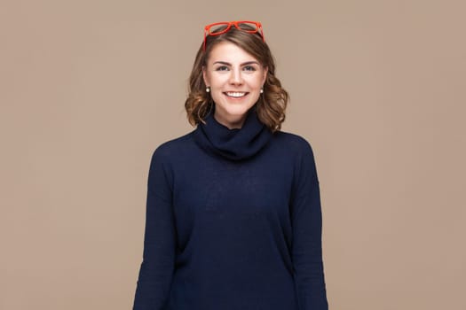 Portrait of positive smiling happy woman with wavy hair in red red glasses on her head looking at camera with satisfied expression. Indoor studio shot isolated on light brown background.