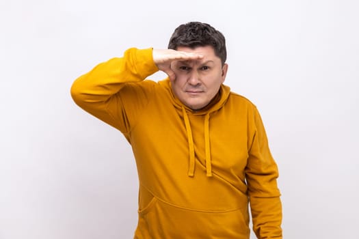 Thoughtful dark haired man looking far away at distance with hand over head, attentively searching for bright future, wearing urban style hoodie. Indoor studio shot isolated on white background.