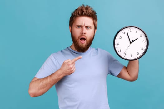 Portrait of shocked astonished surprised handsome bearded man pointing at wall clock, deadline, having puzzled facial expression. Indoor studio shot isolated on blue background.