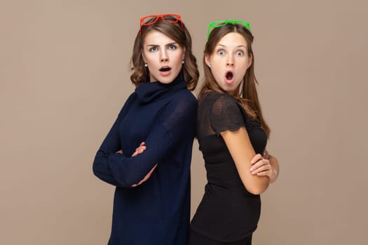 Portrait of two shocked surprised astonished women friends standing back to back and looking at camera with big eyes and open mouth. Indoor studio shot isolated on light brown background.