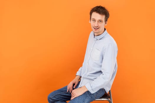 Portrait of handsome attractive man sitting and looking at camera with positive expression, wearing light blue shirt and jeans. Indoor studio shot isolated on orange background.