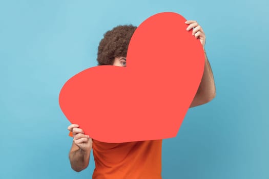 Portrait of man hiding face behind big red heart and looking at camera with curious prying eyes, expressing i love you, feeling affection fondness. Indoor studio shot isolated on blue background.